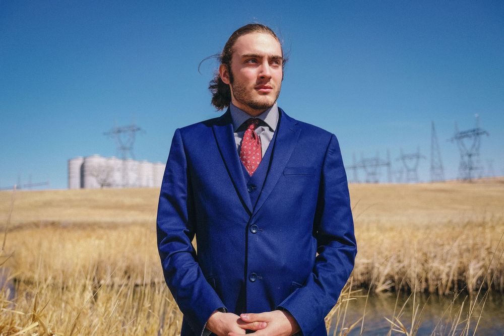 A person in a bright blue suit poses in a golden wheat field against a clear blue sky.