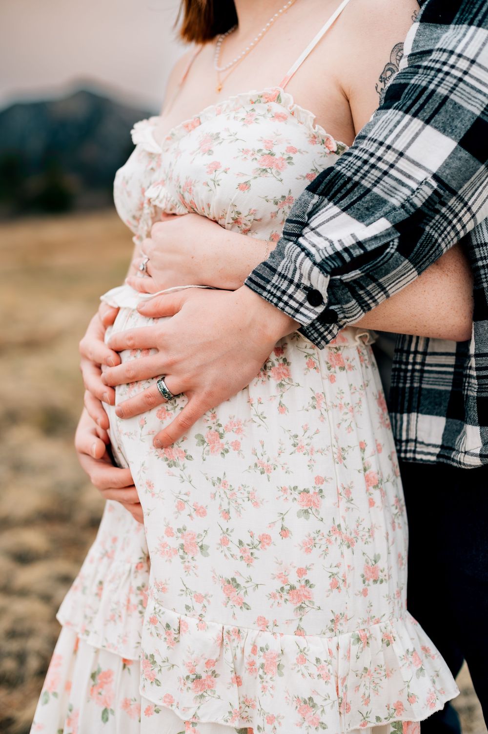 Close-up of embracing couple's hands on belly in floral dress and plaid shirt.