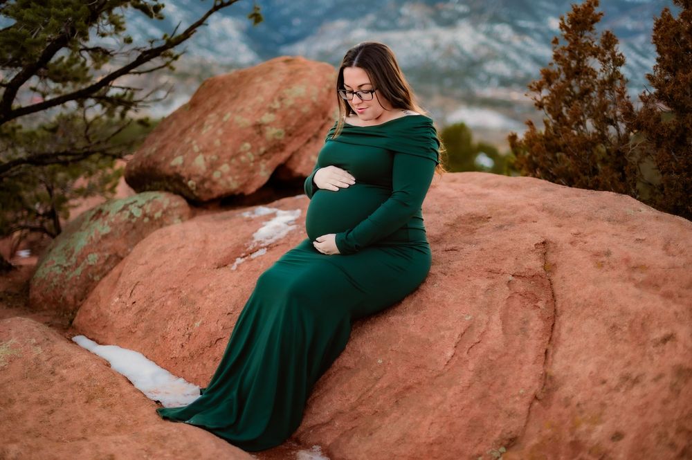 A maternity photo of an expectant mother in emerald dress sitting on red rocks at sunset.