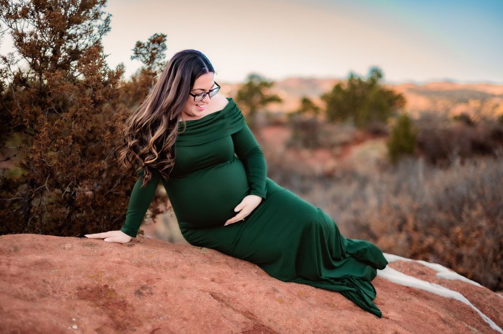 An elegant maternity portrait with a woman in green dress reclining on red rocks outdoors.