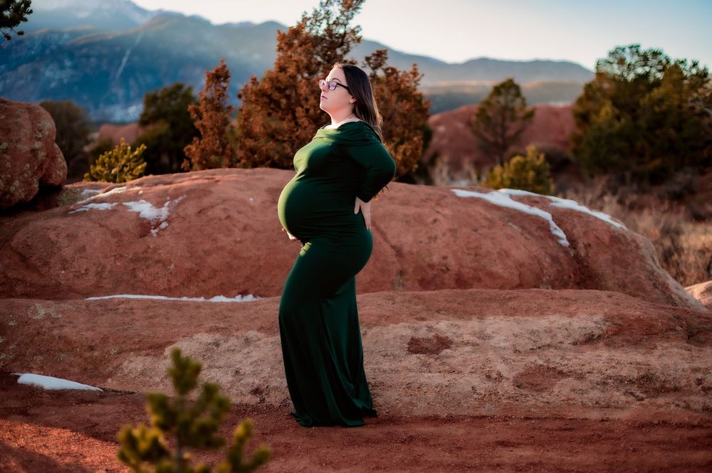 A silhouette of an expecting mother in green dress against mountain landscape at sunset.