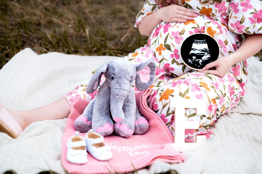 A floral dress and stuffed elephant toys arranged on a white blanket for a maternity photoshoot.