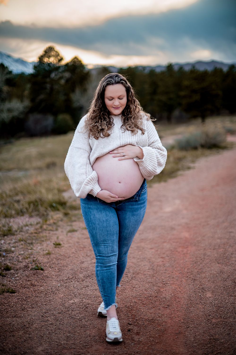 An expectant mother in a white sweater walks along a dirt path during sunset in a mountain setting.