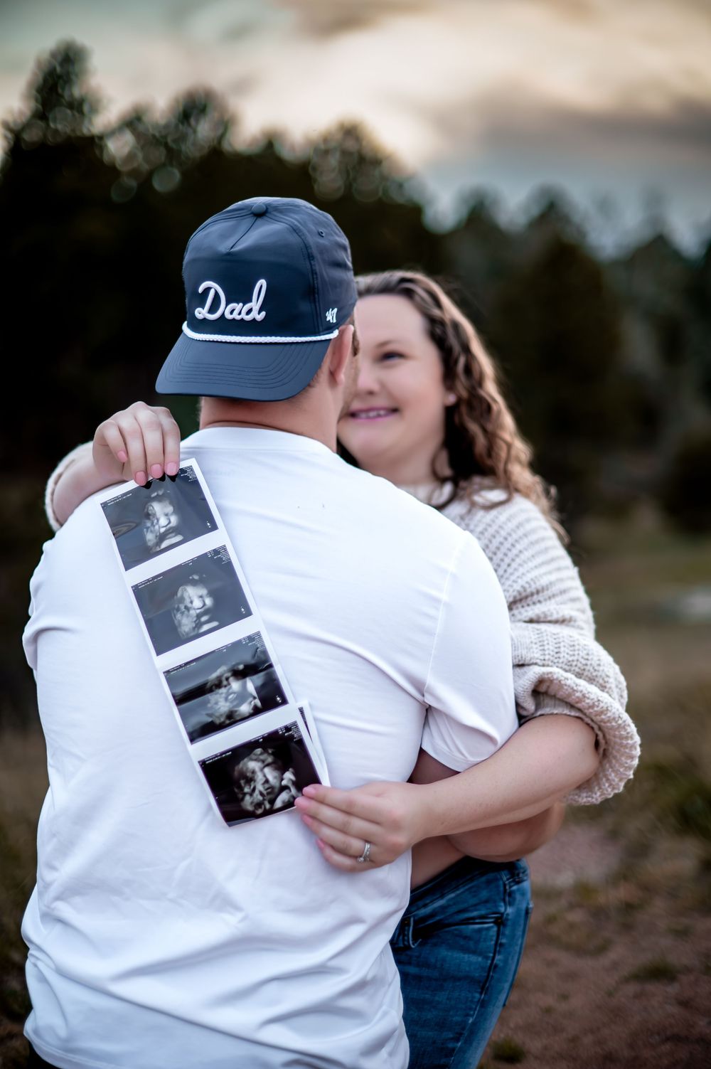 A couple shares an intimate moment while looking at ultrasound photos outdoors during sunset.
