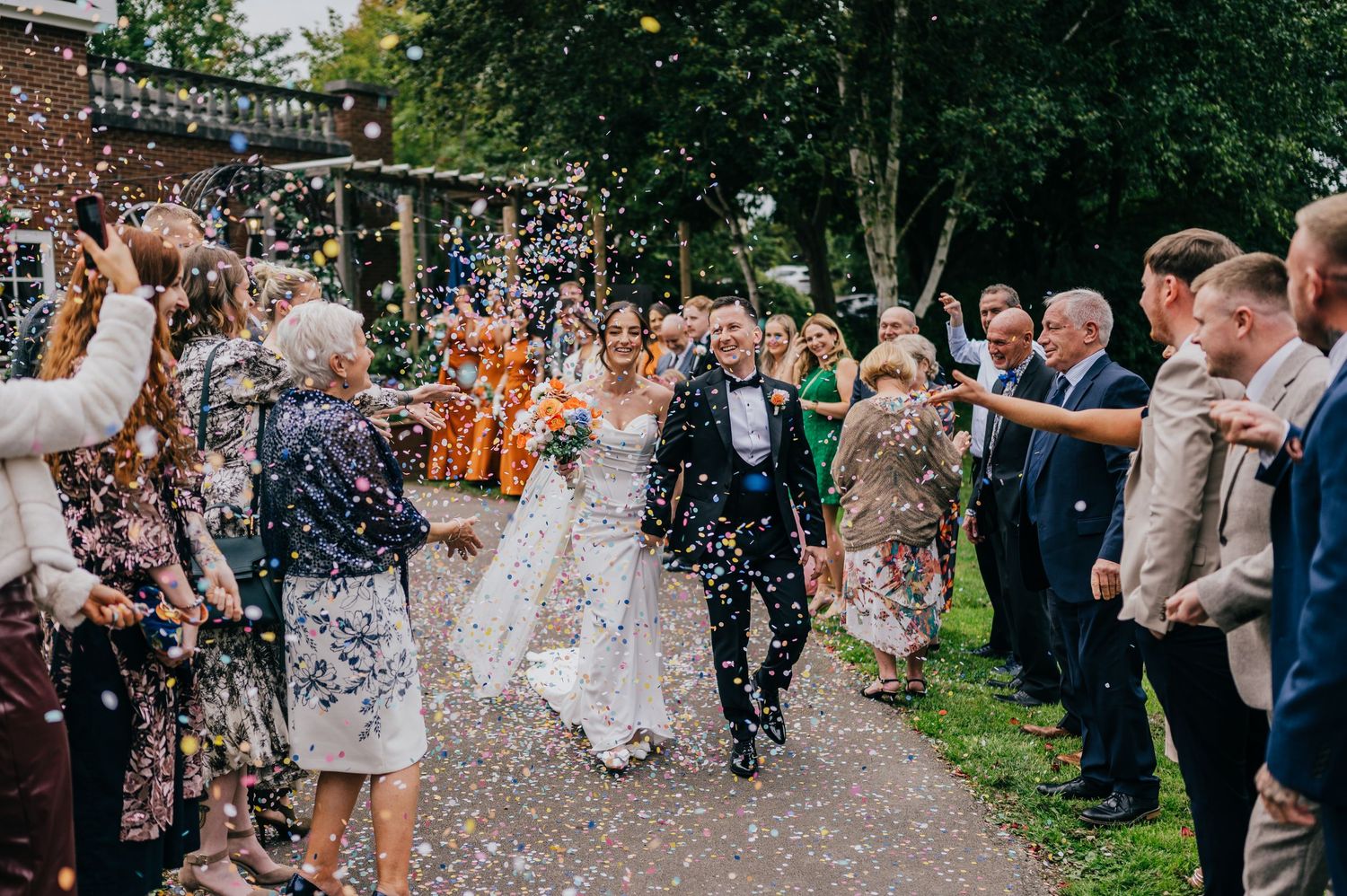 Bride and groom walk through confetti shower surrounded by cheering wedding guests on a tree-lined path.