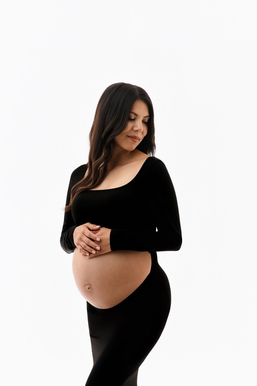 Elegant pregnant woman in black long-sleeve outfit holds her bare baby bump against white background.