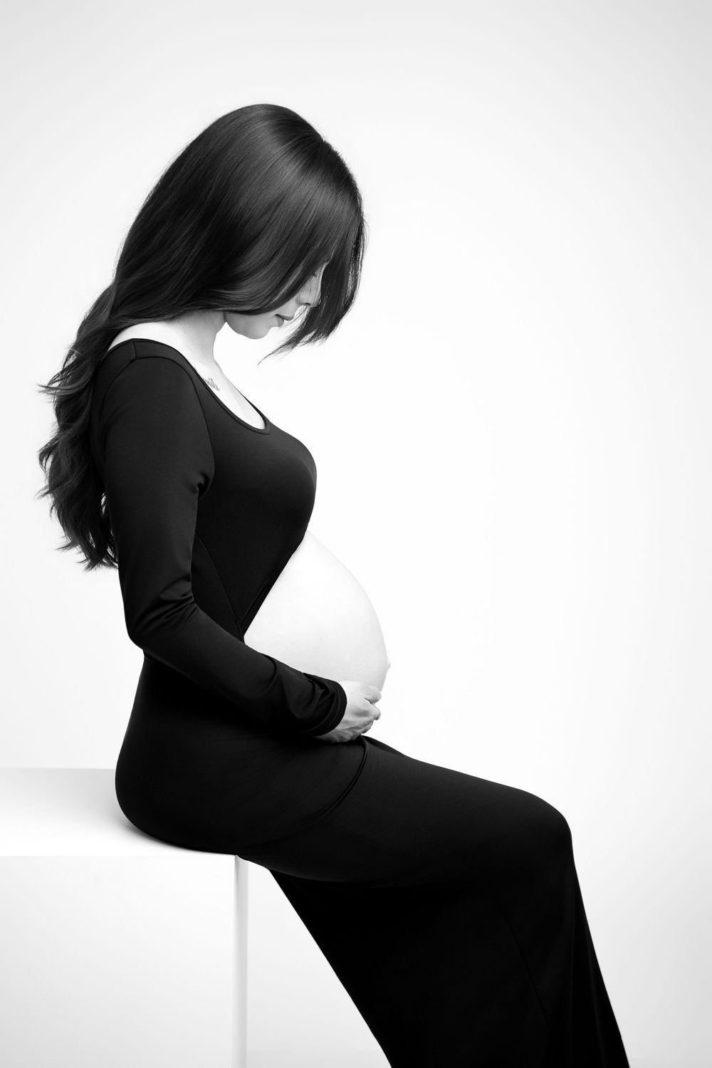 Black and white portrait of pregnant woman in black dress sitting on cube holding her baby bump.
