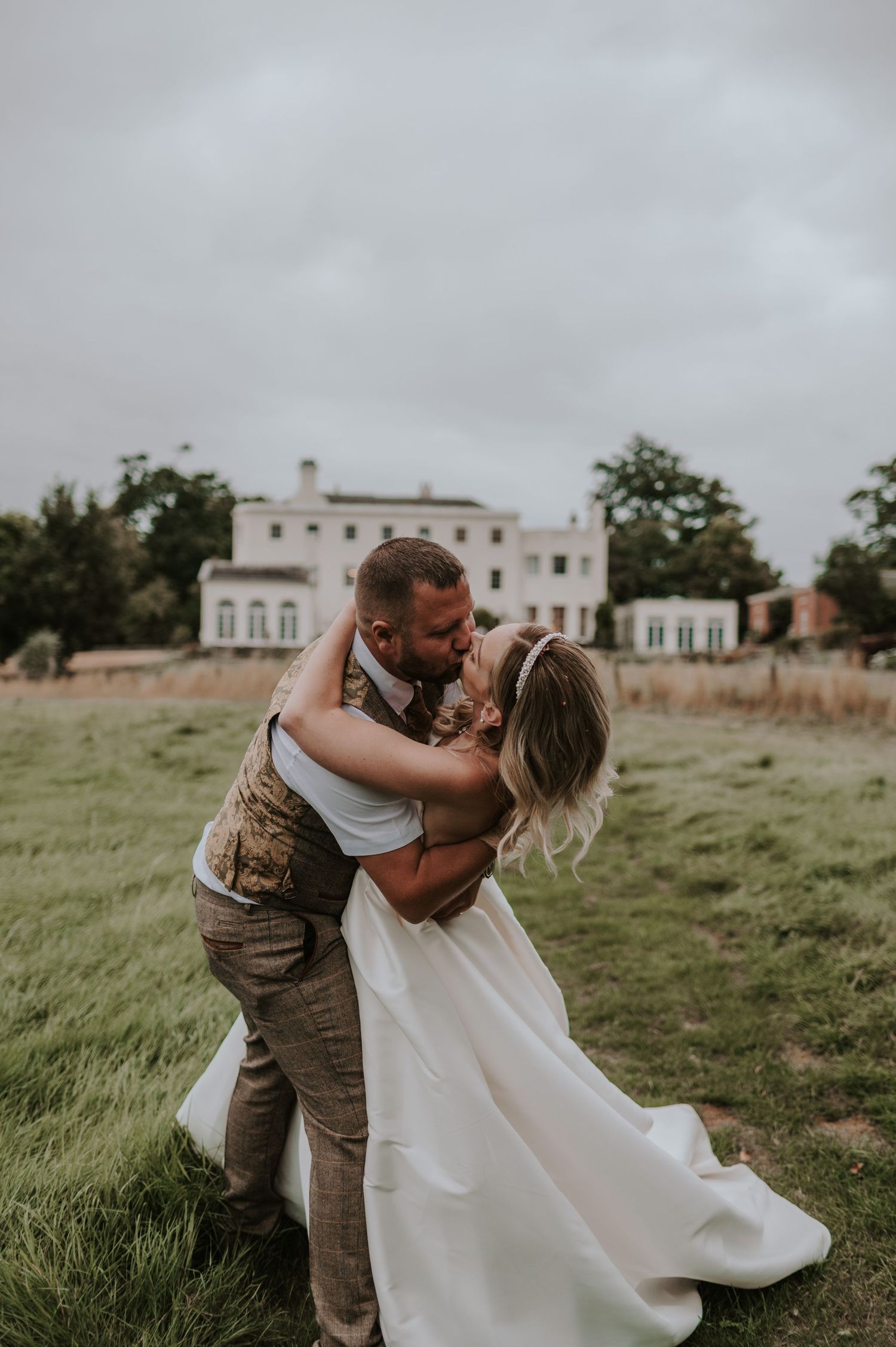 Couple embraces in romantic spin pose on grassy lawn with elegant Rockbeare Manor in the background.