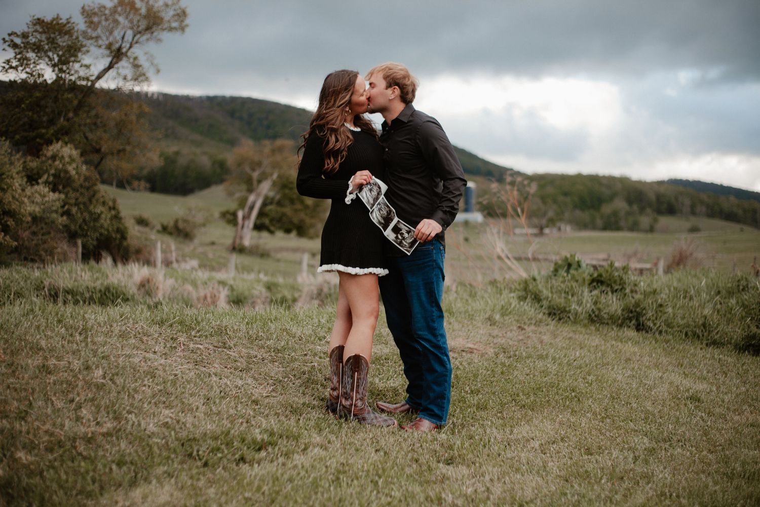 Couple kissing in a grassy field with rolling hills, woman in black dress holding ultrasound photos.