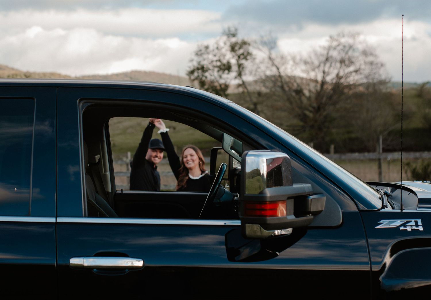 Couple celebrating joyfully inside a black truck with arms raised, scenic hills in background.