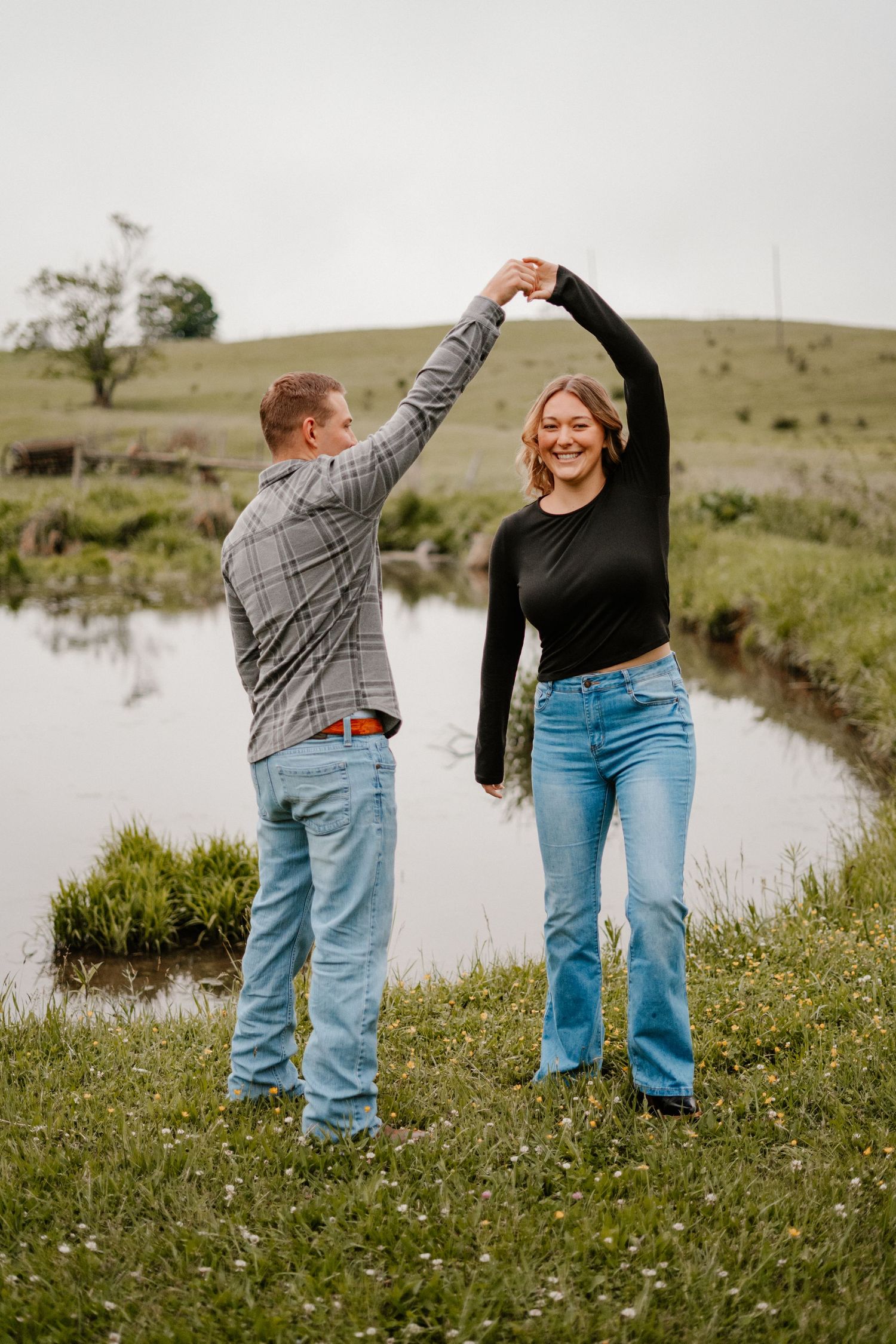 Couple dancing by a reflective pond on a green hillside, woman smiling in jeans and black top.