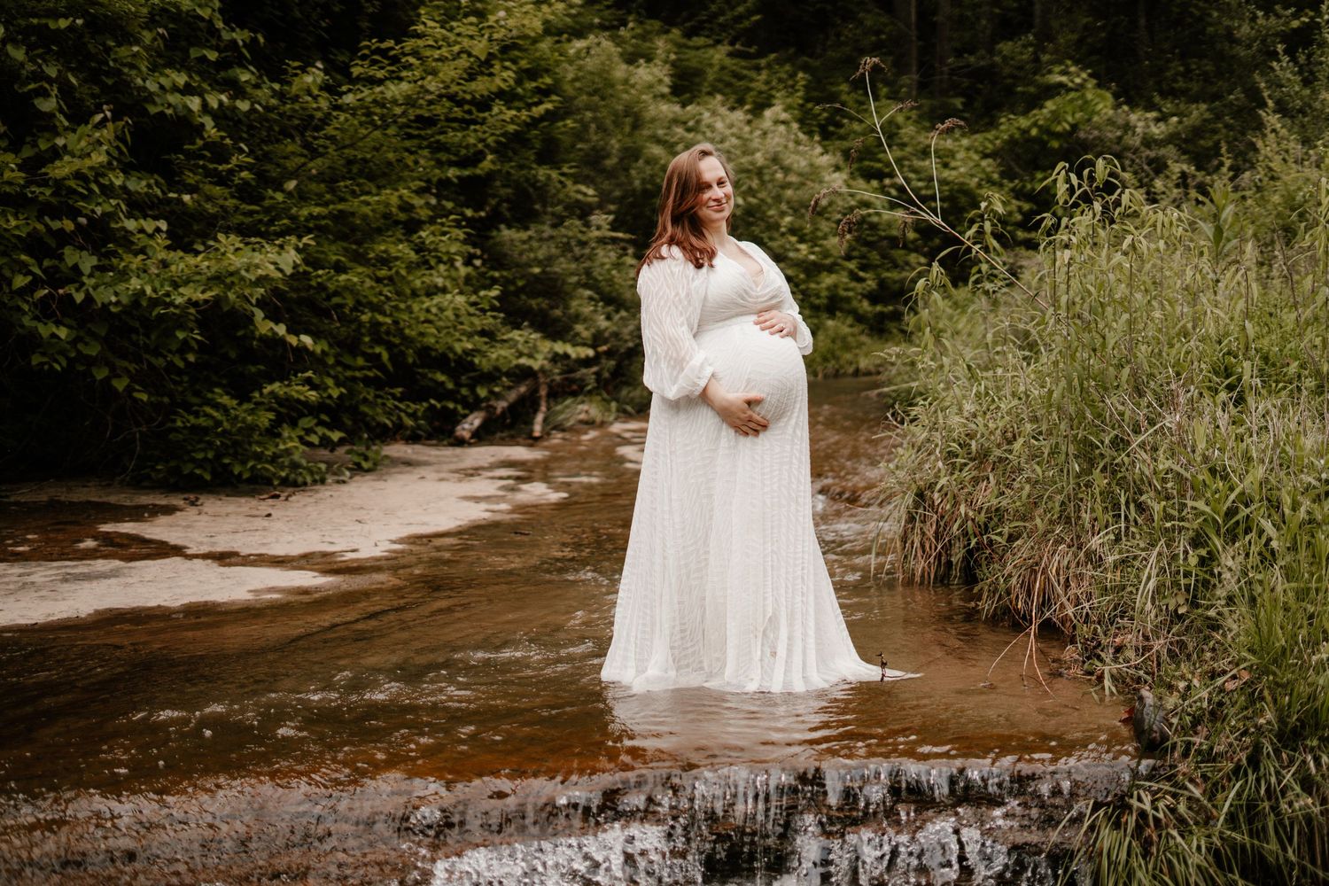 Pregnant woman in white gown standing in a shallow creek surrounded by lush green forest.
