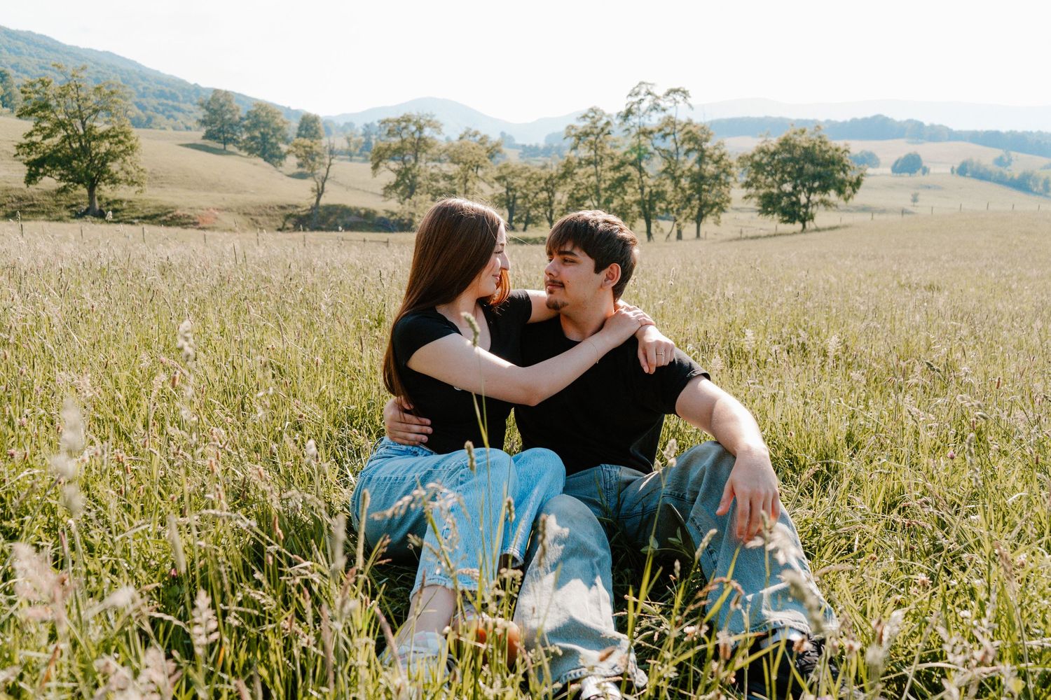 Young couple sitting closely together in a lush green meadow with rolling hills in the background.