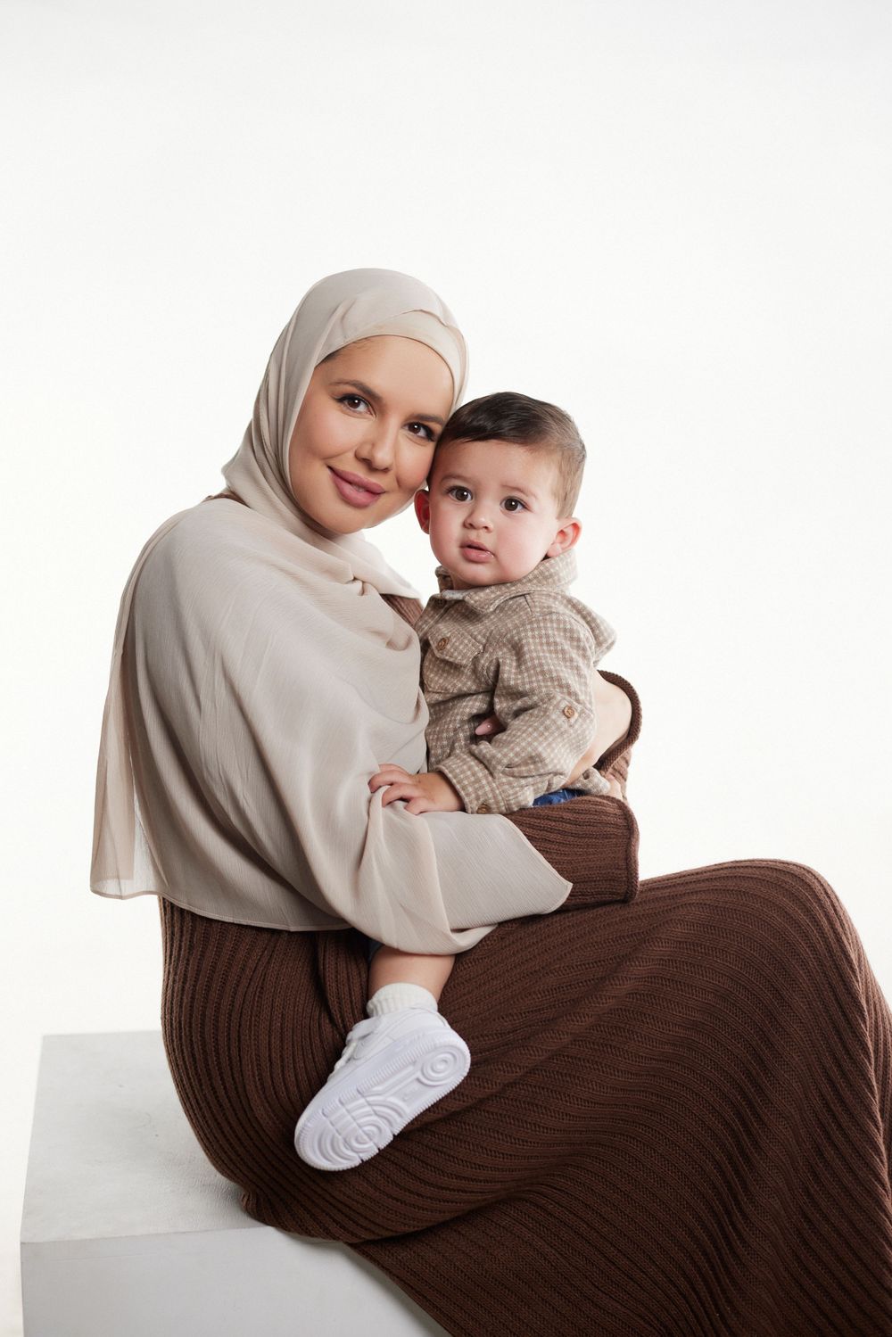 Mother in beige hijab holding baby wearing brown sweater on white background.