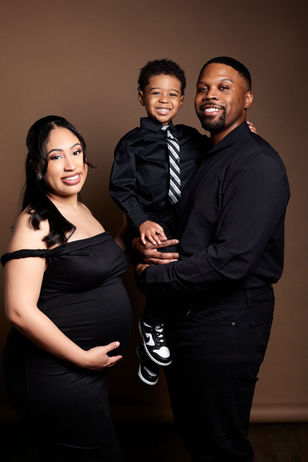 Elegant family portrait session with coordinated black outfits against a brown backdrop.