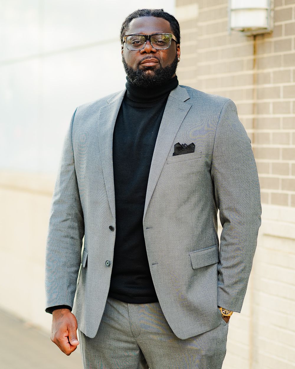 Professional wearing a gray suit jacket with black turtleneck stands against beige wall.
