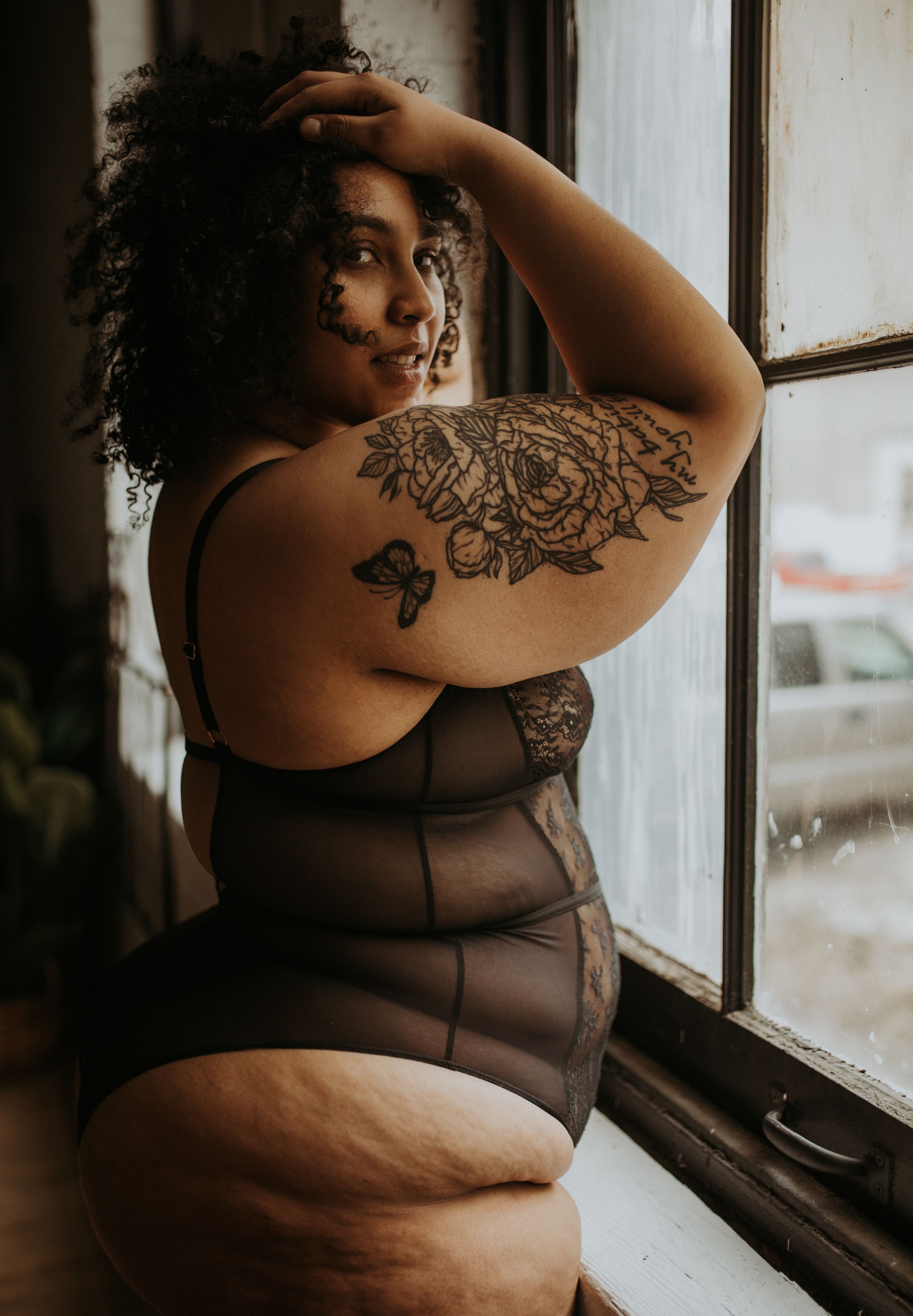 Artistic boudoir photo of person in sheer black lingerie posing by window with intricate floral tattoo on shoulder.