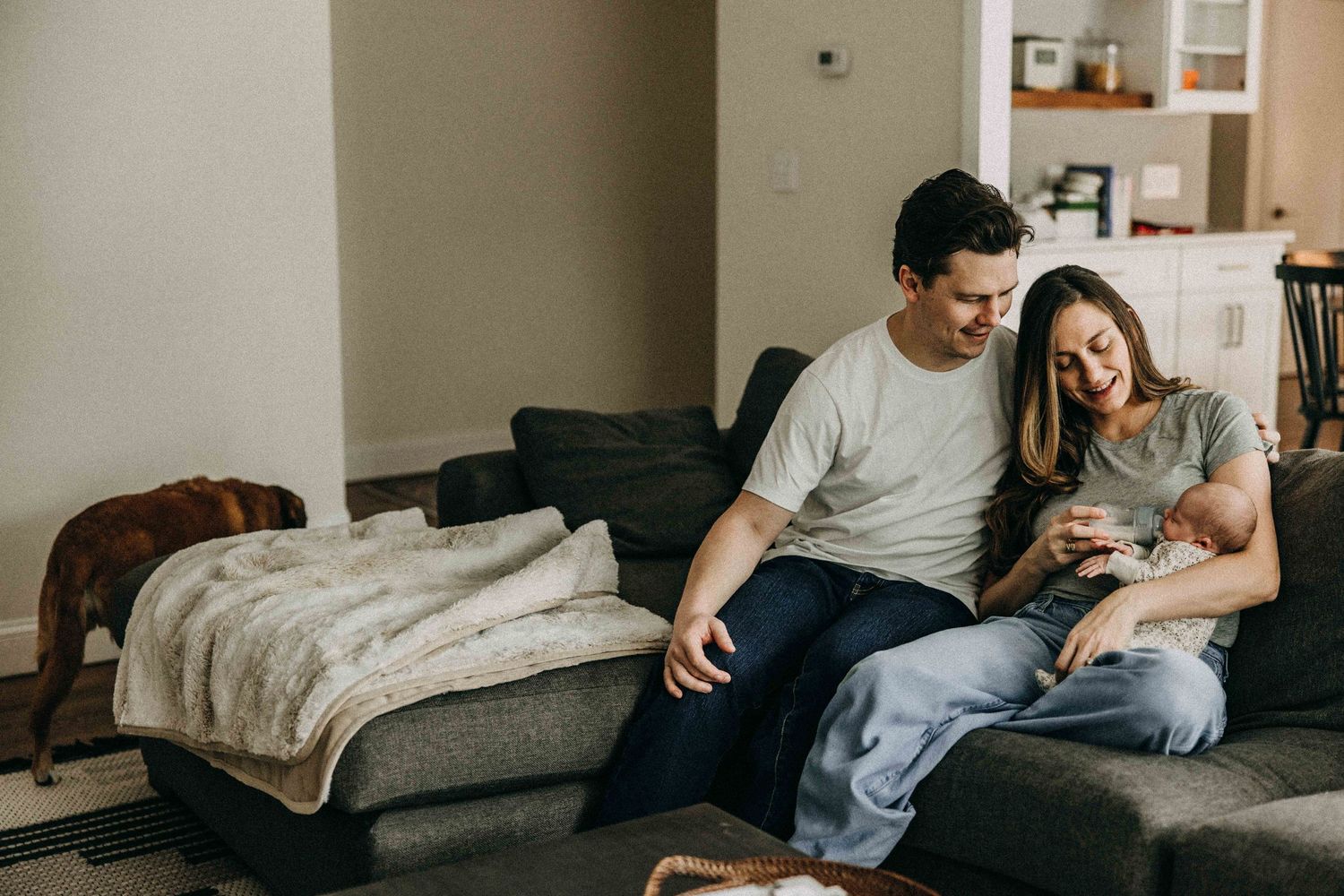 A young couple shares a tender moment with their newborn on a grey couch.