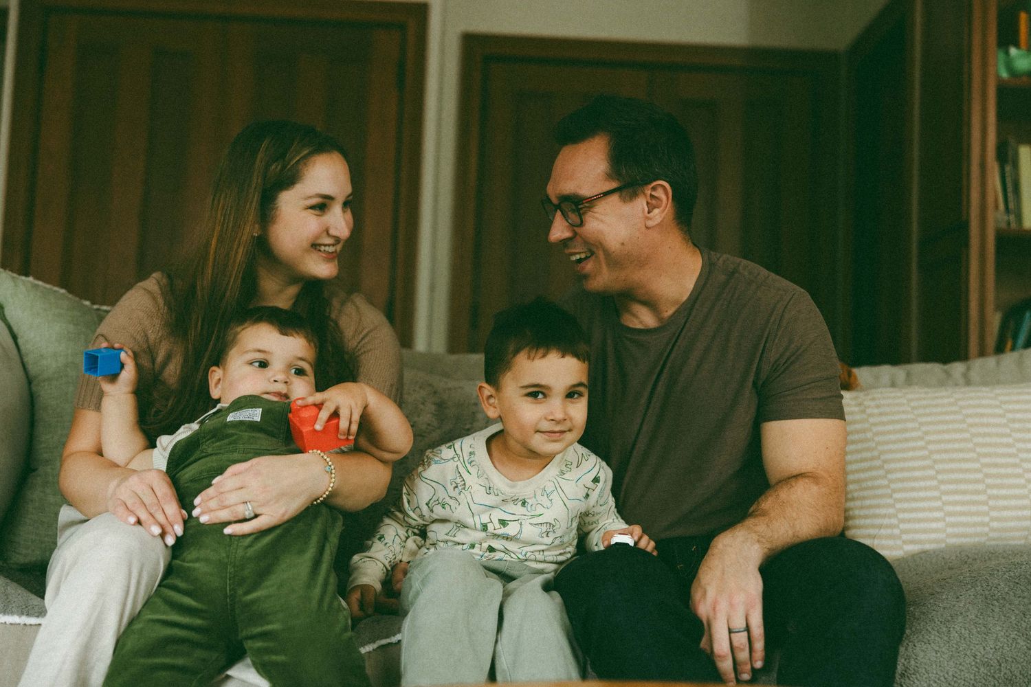 A happy family of four sits together on a couch, with two young children and smiling parents in a cozy living room.