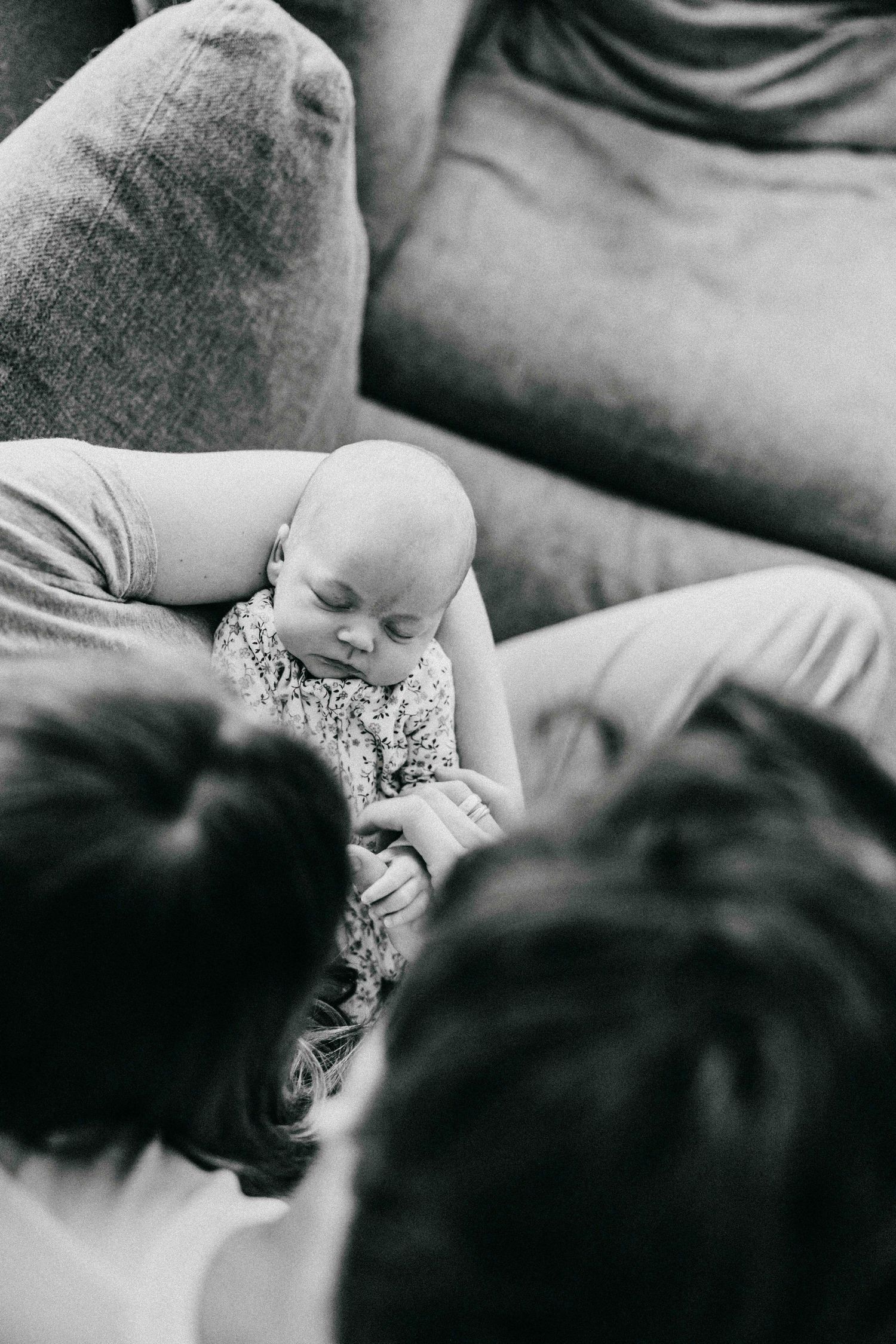 Black and white photograph of a sleeping baby in a floral outfit laying between parents on a couch.