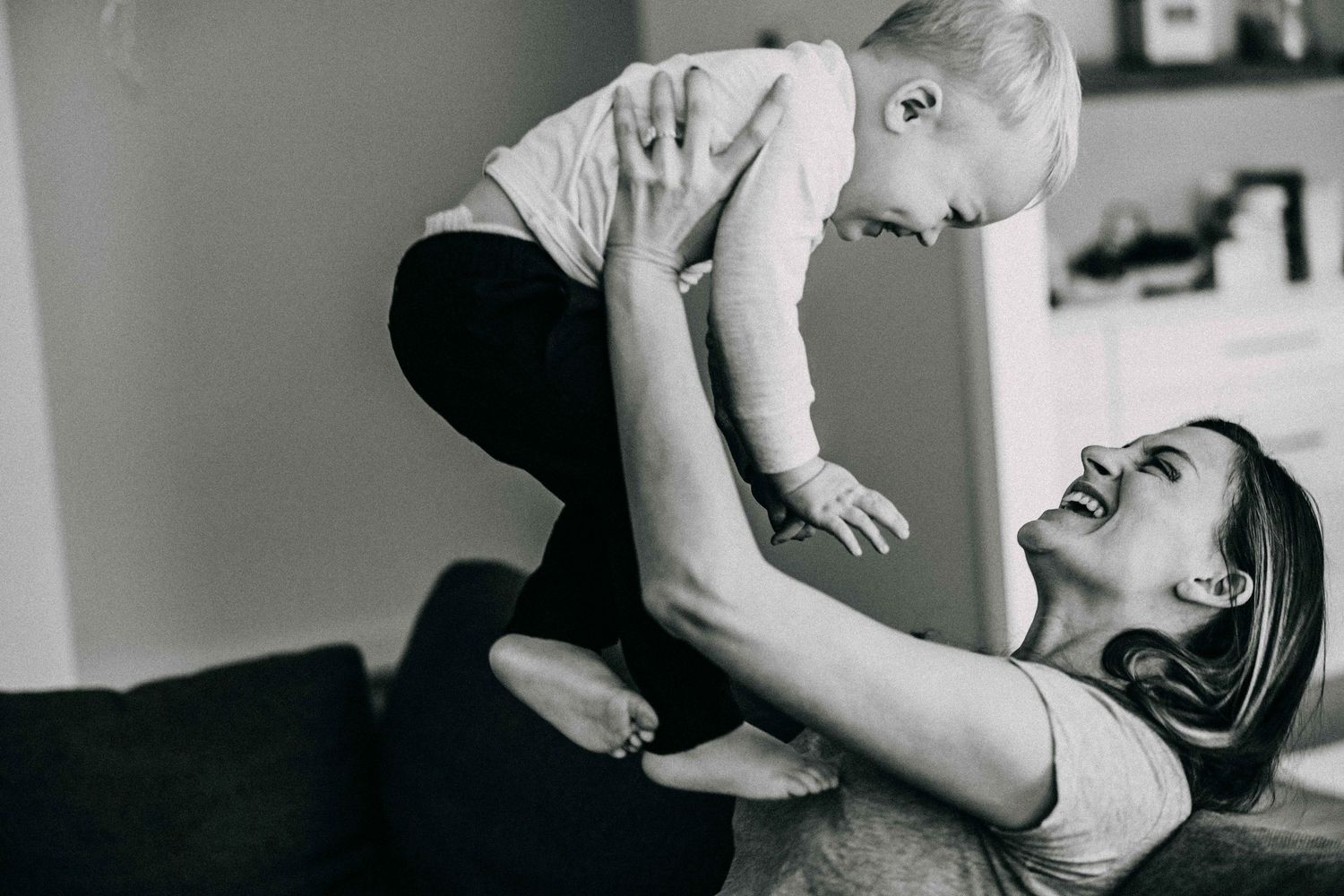 Parent lifts baby up high during joyful playtime on the sofa in black and white photo.