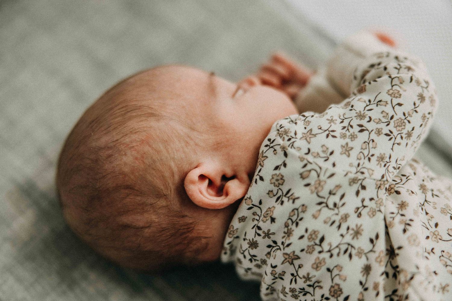 Close-up profile of a sleeping infant in a floral pattern onesie resting on grey fabric.