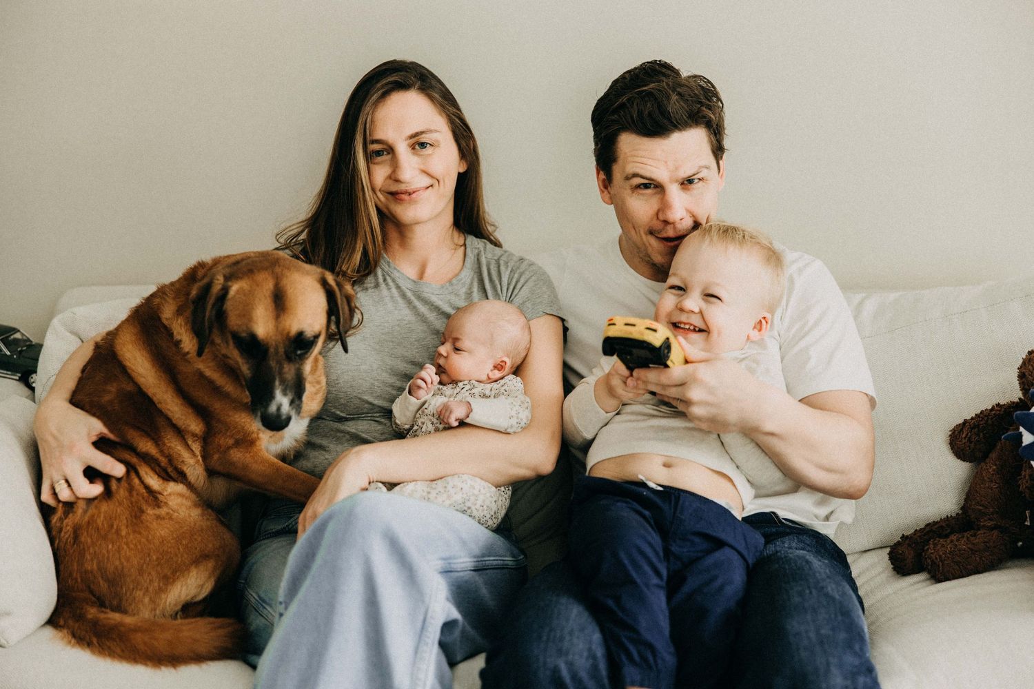 Couple sits on couch with their dog and infant twins in cozy home setting.