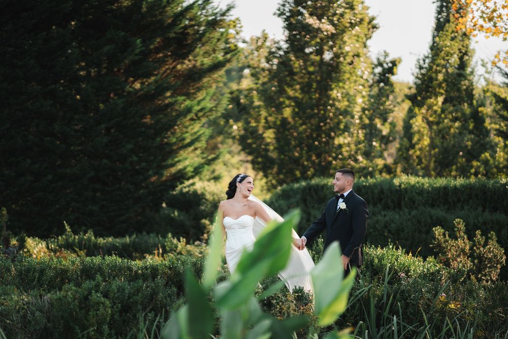 A couple in formal wedding attire stands in a lush garden setting during golden hour sunlight.