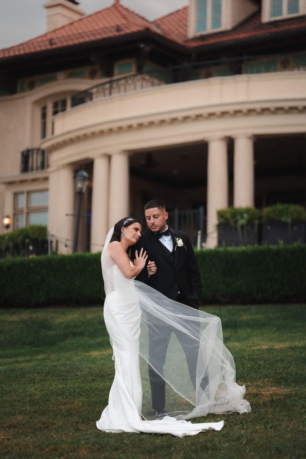 A bride and groom share an intimate moment on a grassy lawn with her veil flowing in the breeze.