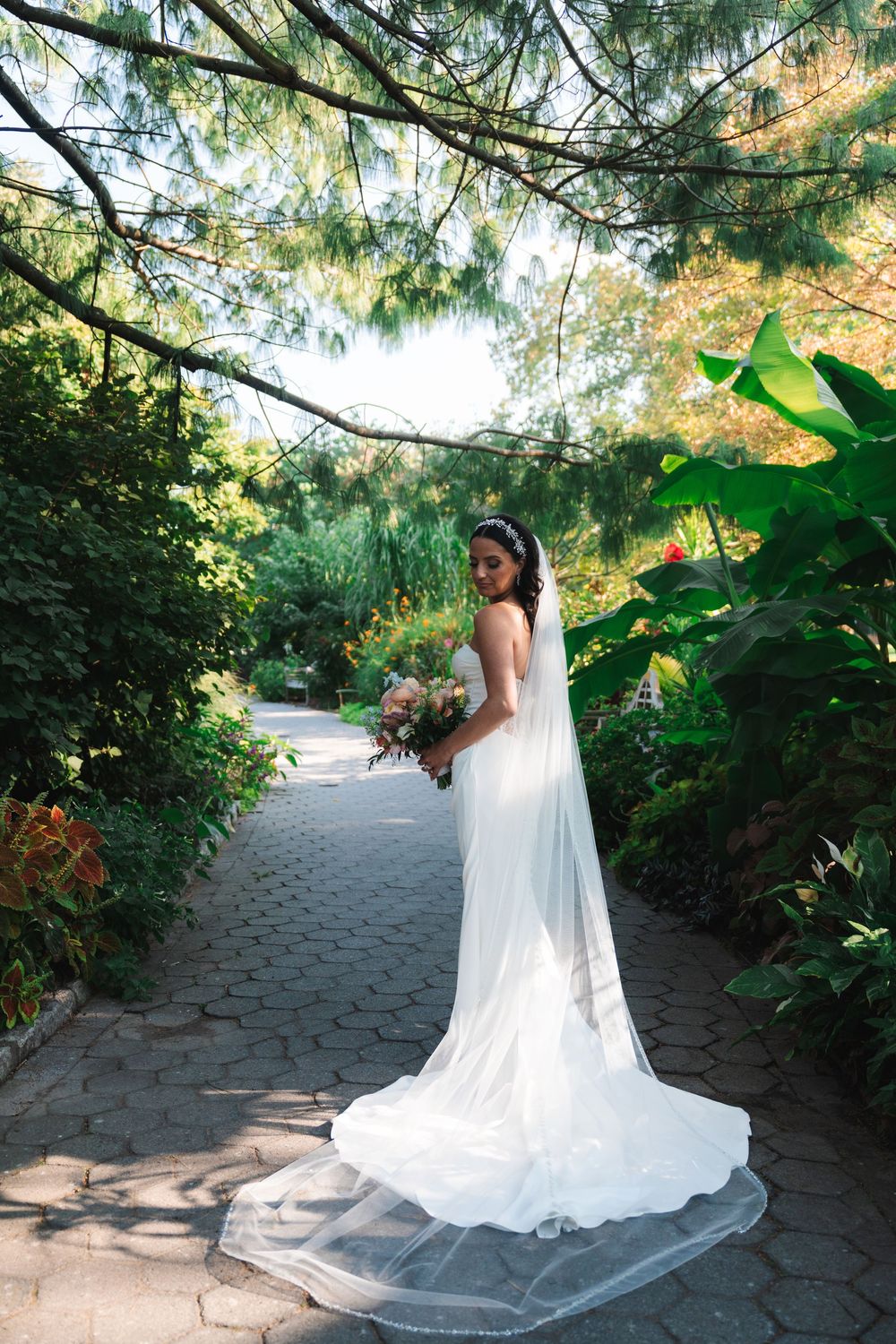 A bride in a flowing white wedding gown stands on a garden path surrounded by lush greenery on a sunny day.