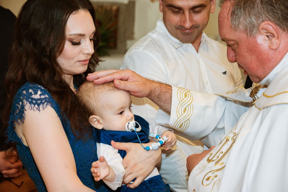A priest performs a baptism ceremony for a baby wearing blue, held by someone in a navy dress during a church service.