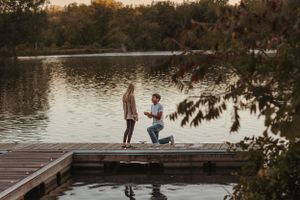 A romantic proposal moment unfolds on a wooden dock by a serene lake at sunset.