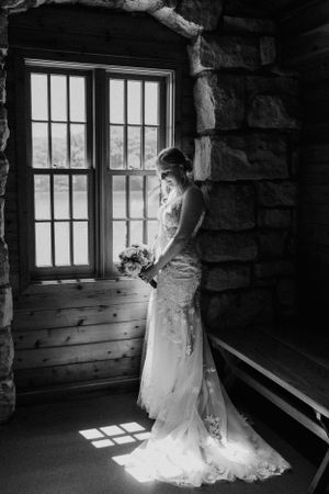 A bride in lace gown stands by a window in dramatic black and white lighting.