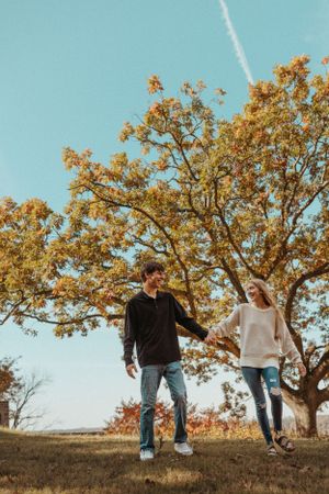 A couple holding hands while walking on a hill with autumn trees and blue sky.