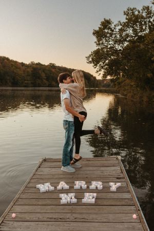 A couple embraces on a dock with 'Marry Me' spelled out in letters.