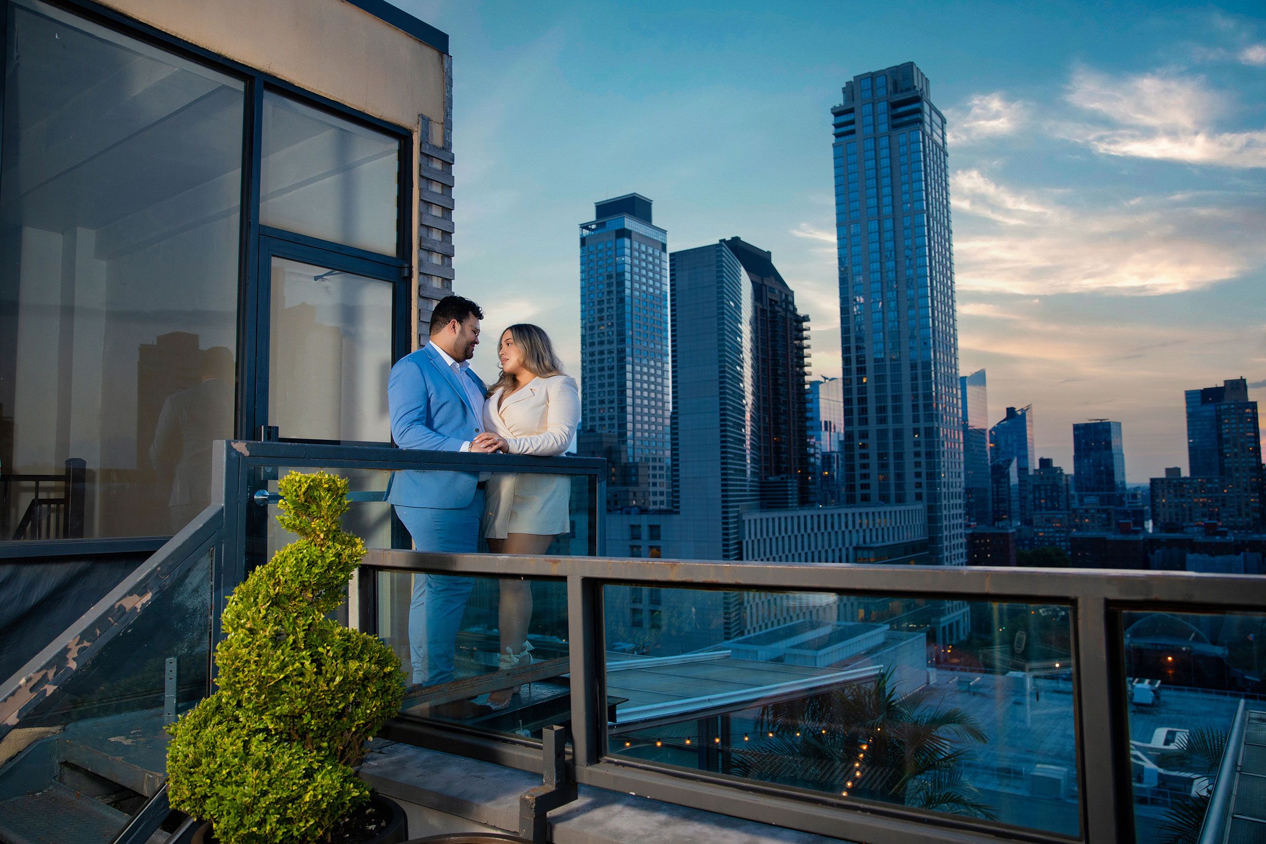 Two people share a romantic moment on a luxury high-rise balcony overlooking a dramatic cityscape at dusk.