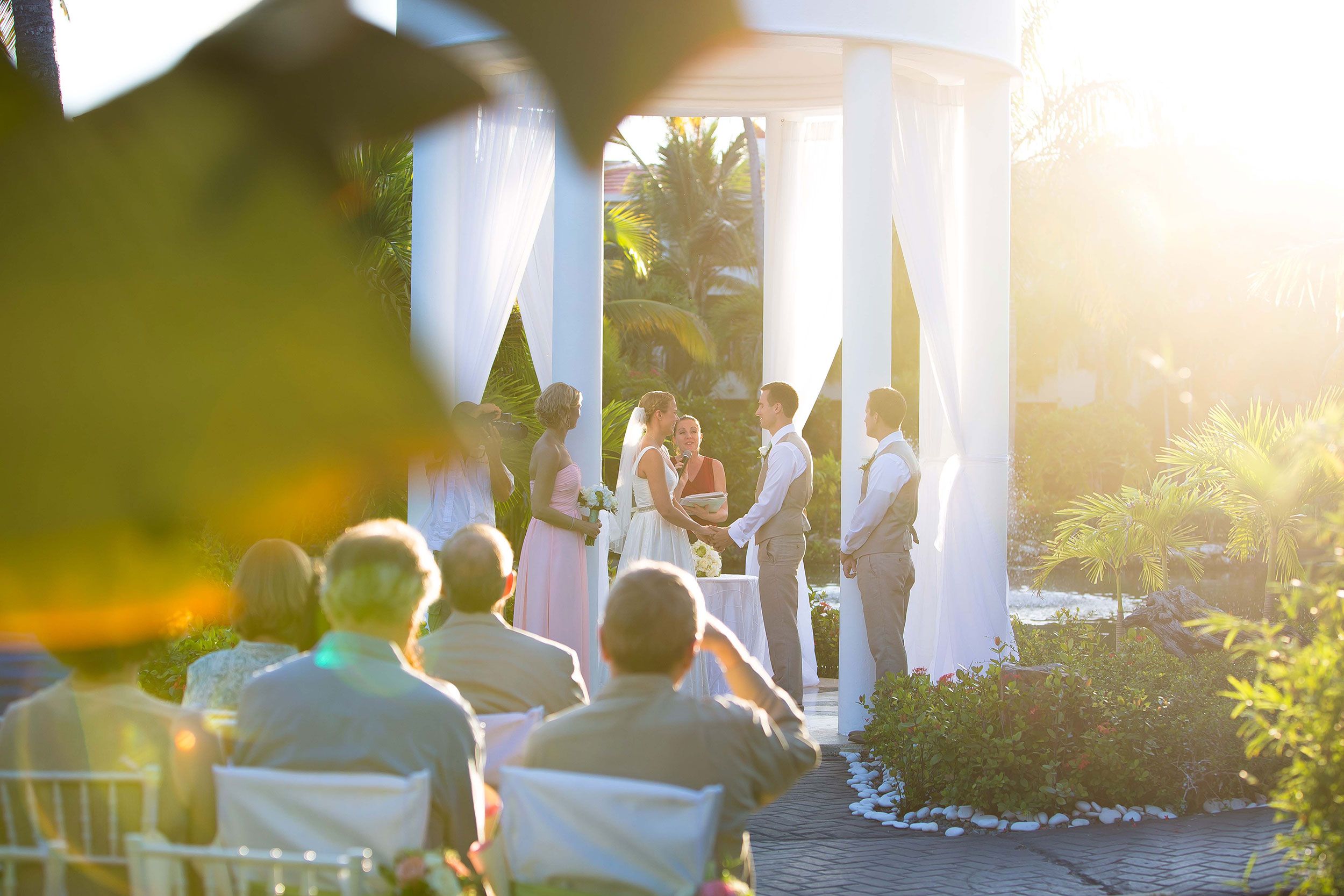 Outdoor wedding ceremony takes place in sunlit white gazebo with guests seated in garden setting.