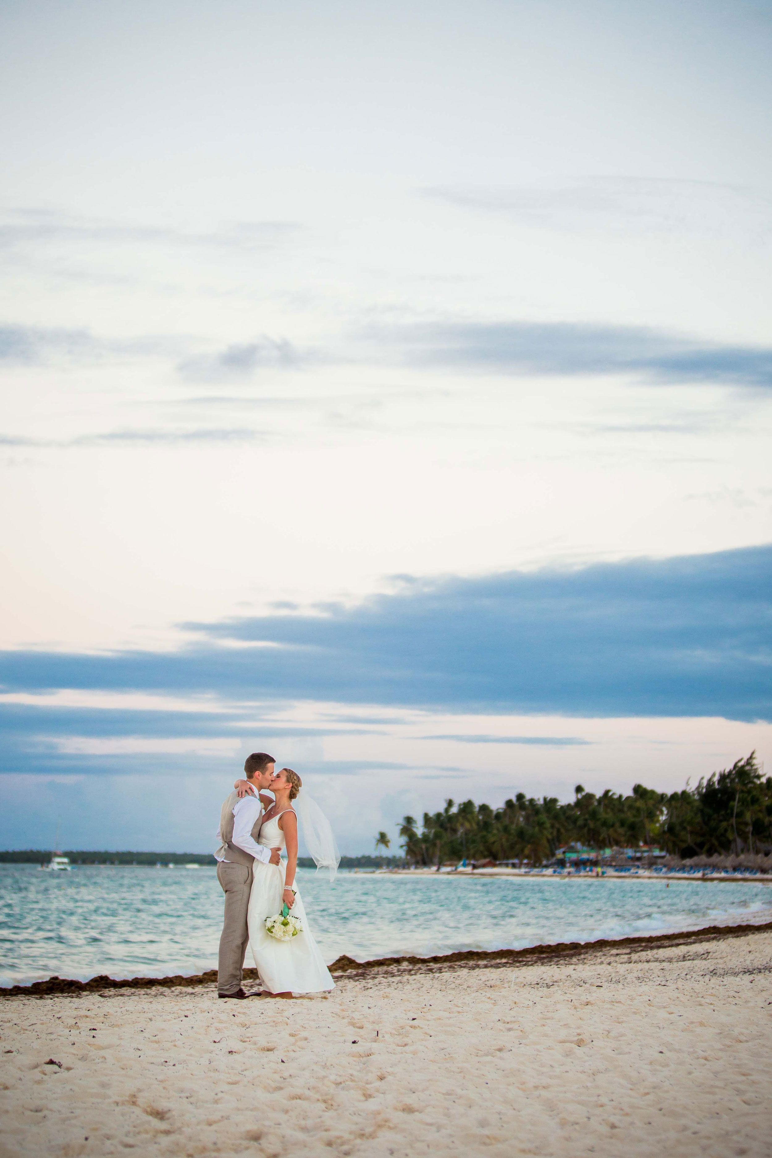 A romantic beach wedding couple shares an intimate moment at sunset on a tropical shoreline with palm trees in the distance.
