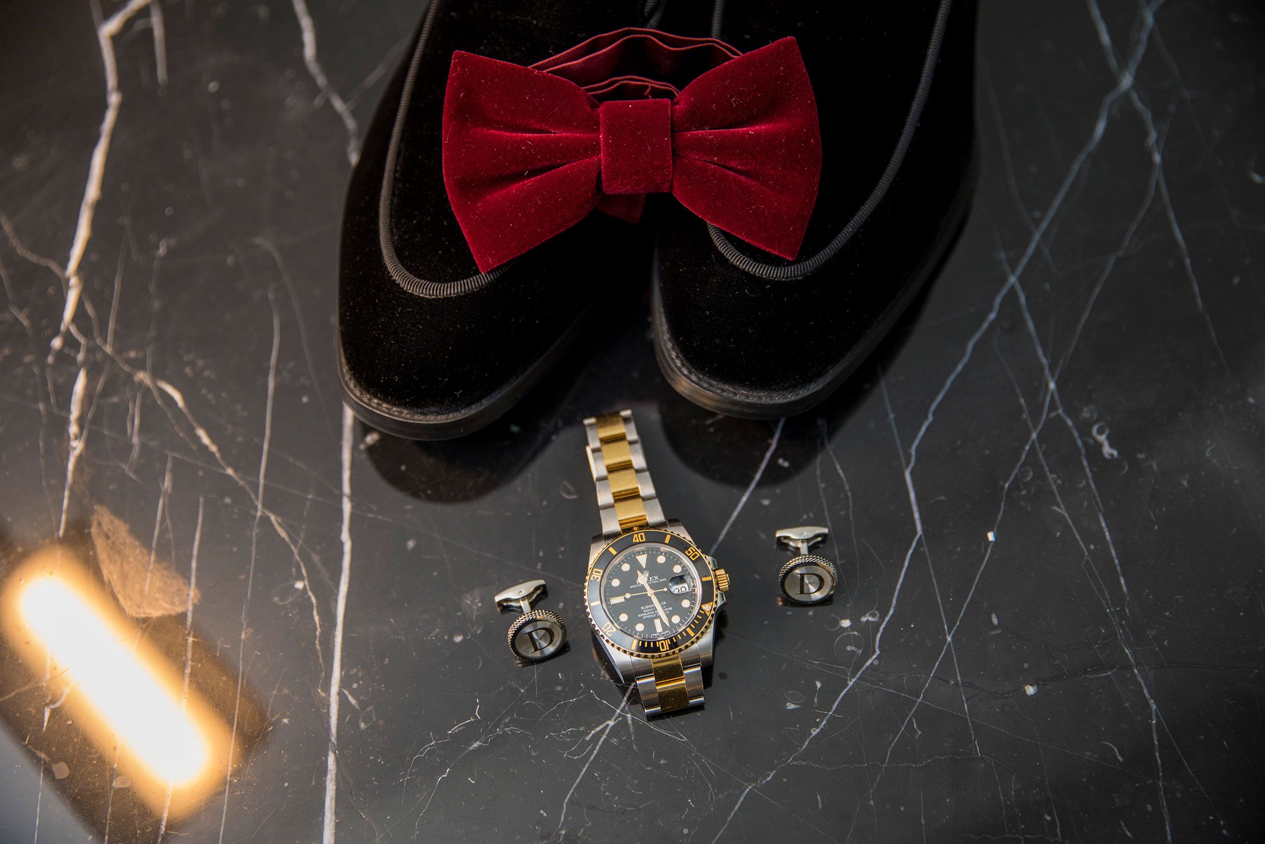 Elegant groom's accessories featuring a red velvet bow tie, black dress shoes and gold watch on dark surface.