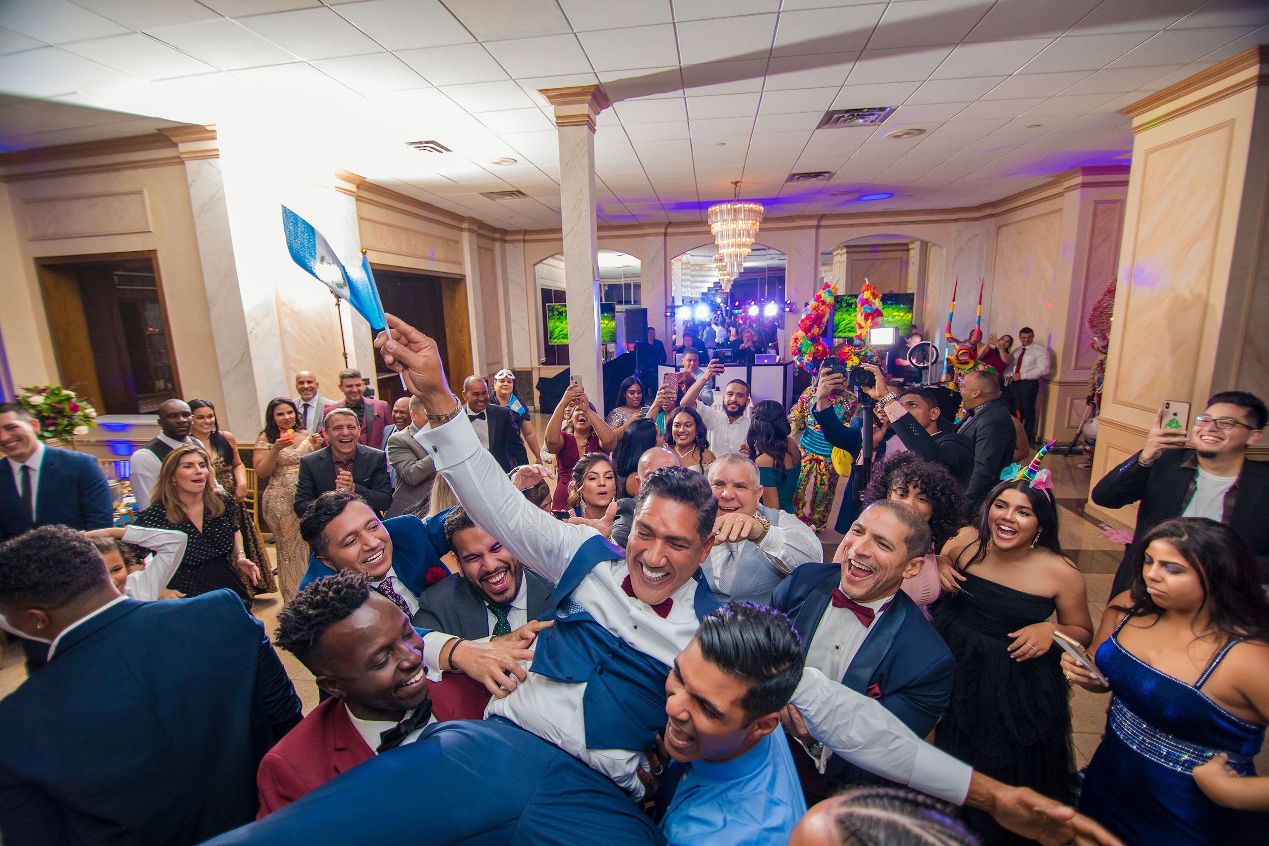 Large group celebrates at wedding reception with people dancing and holding flags.