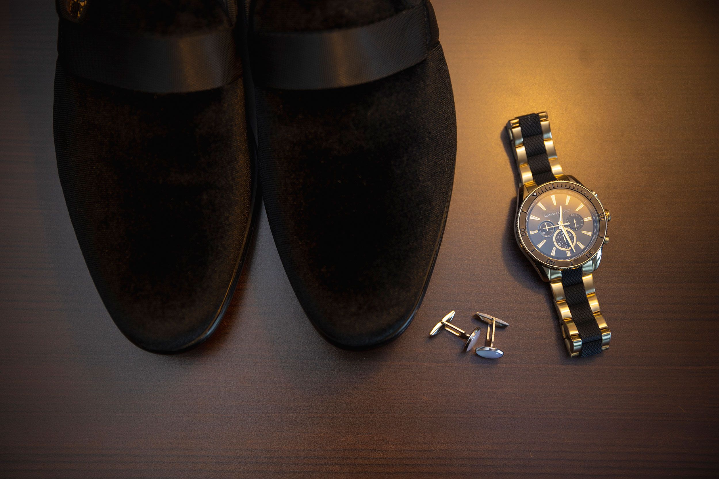 Black suede loafers arranged next to a luxury wristwatch and elegant cufflinks on dark wooden surface.