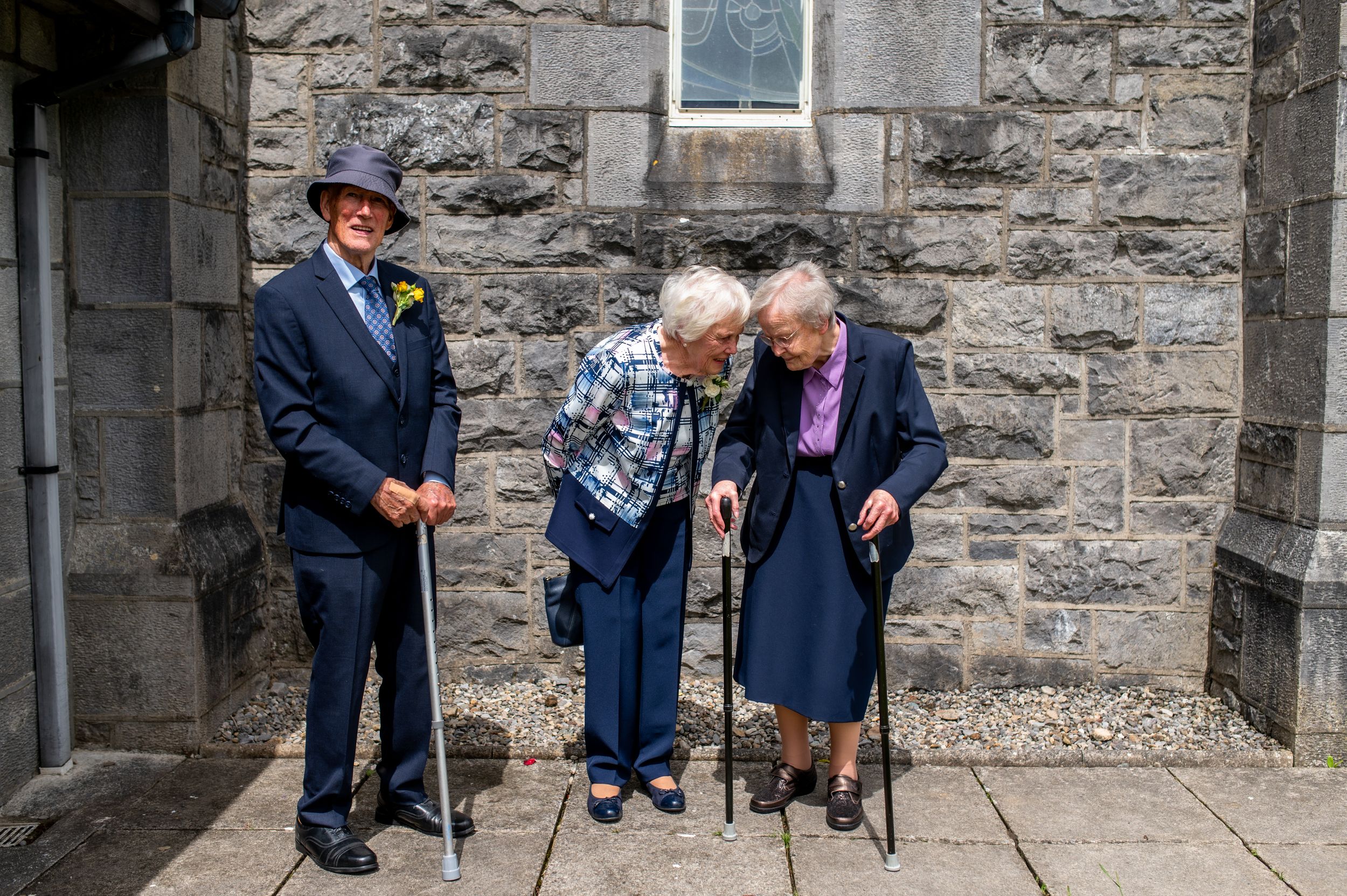 Three people stand together against a stone church wall on a sunny day.