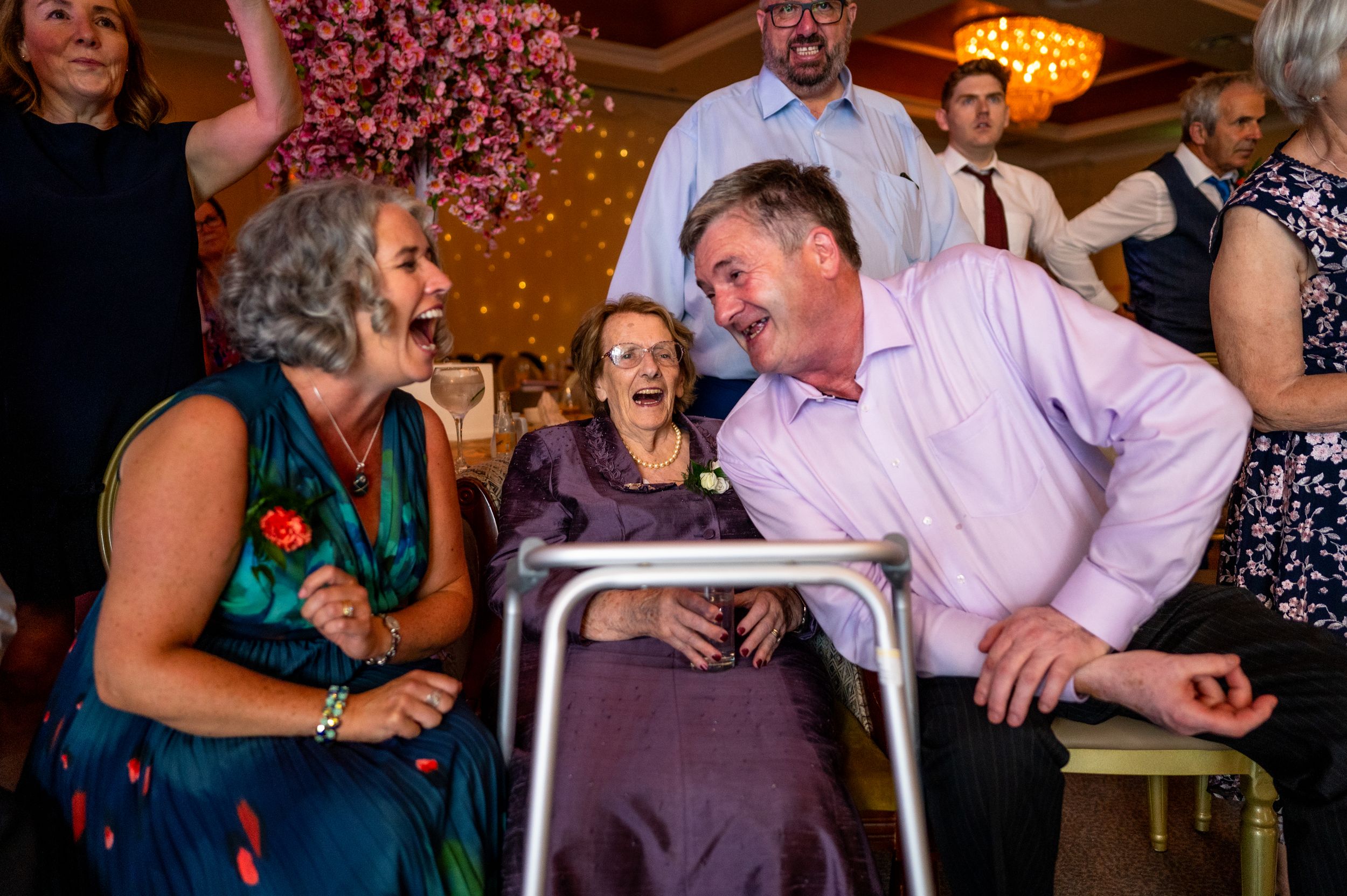 A group of friends laugh and dance together at a dimly lit party or reception venue with warm lighting.