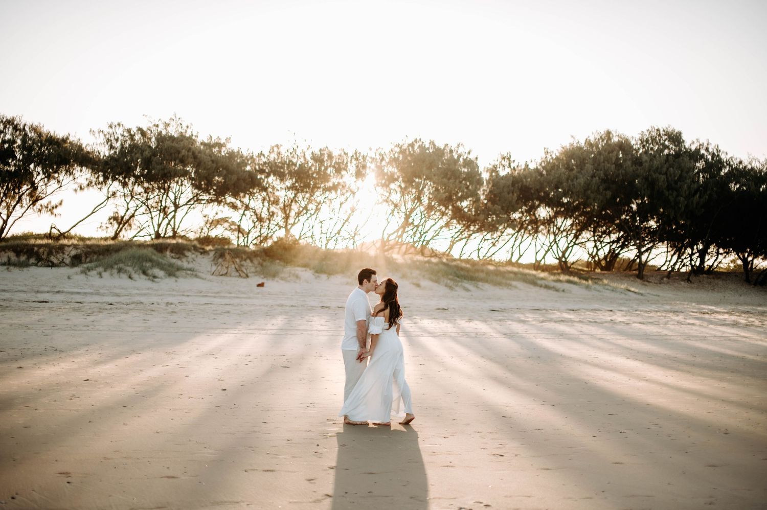 A couple walks through a sunlit pine forest in wedding attire.