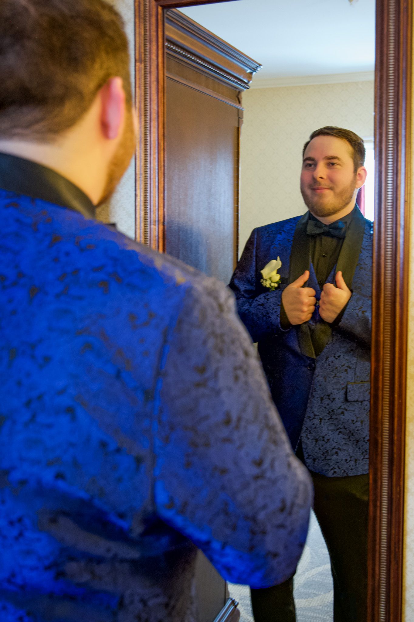 Confident young man in blue floral tuxedo giving thumbs up while checking his look in mirror.