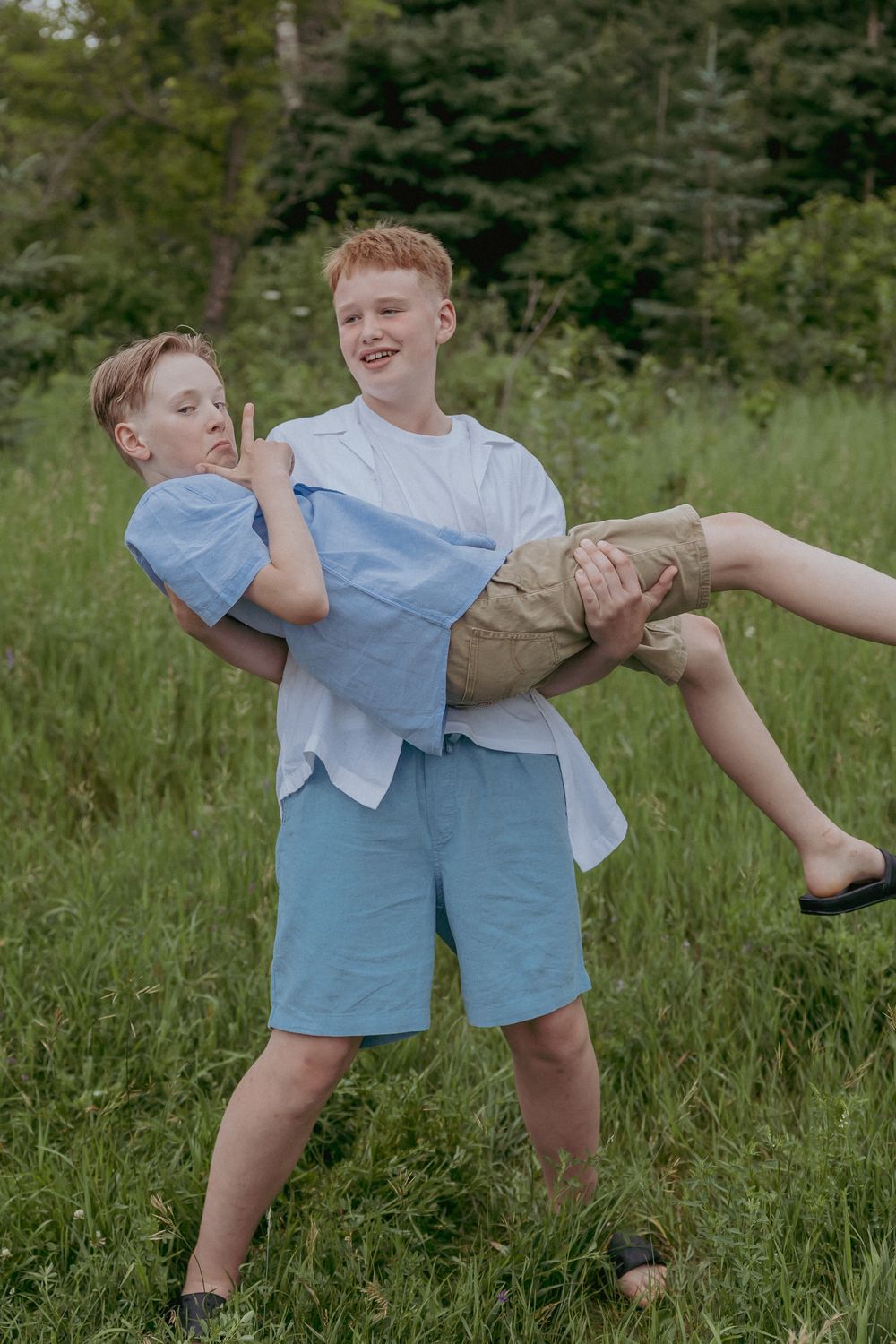 Two red-haired boys playing outdoors, one carrying the other in his arms while laughing in a green field.