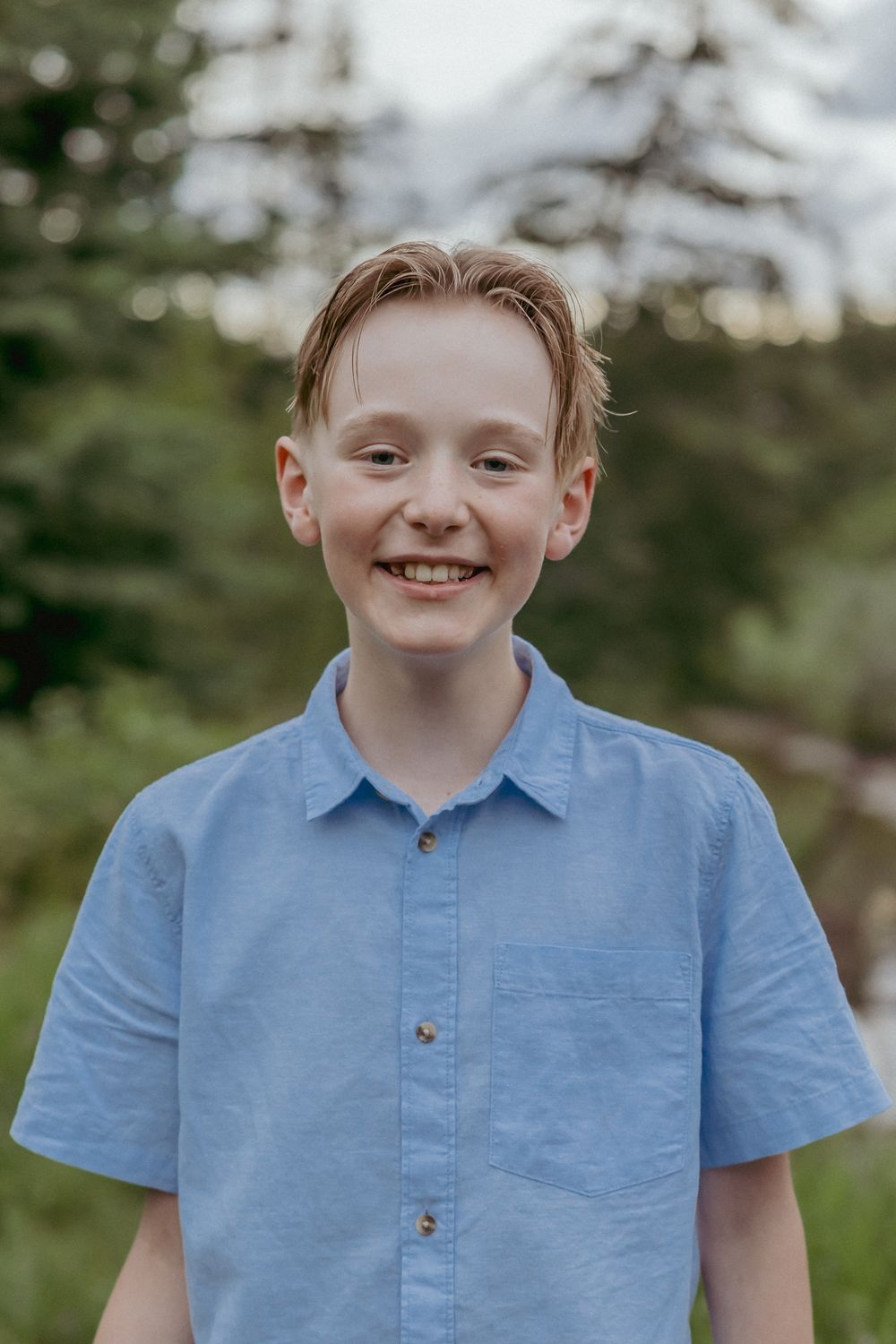Smiling young boy with red hair wearing a light blue button-up shirt outdoors with green trees in background.