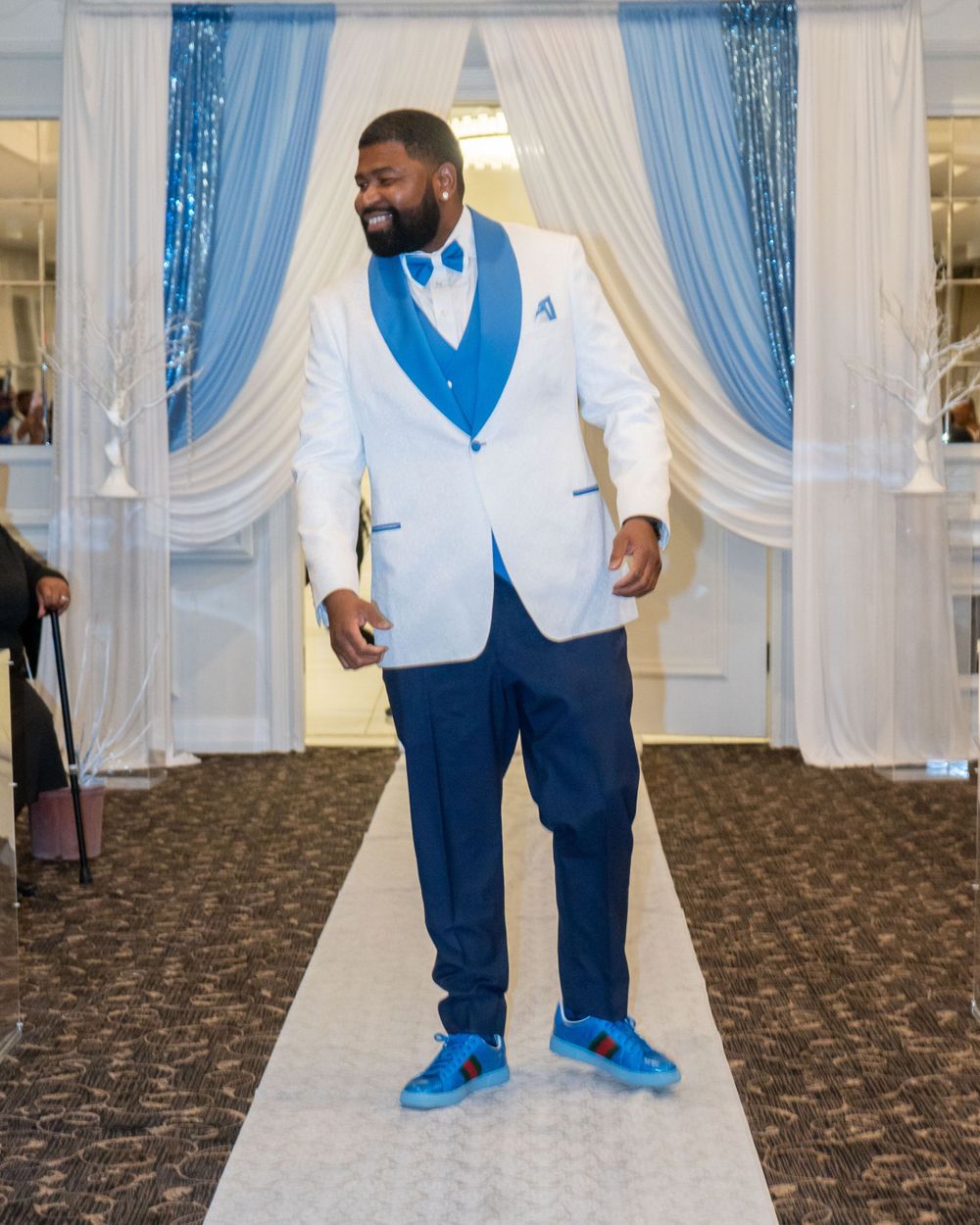 Groom wearing white jacket with blue vest and bow tie walking down white aisle runner.