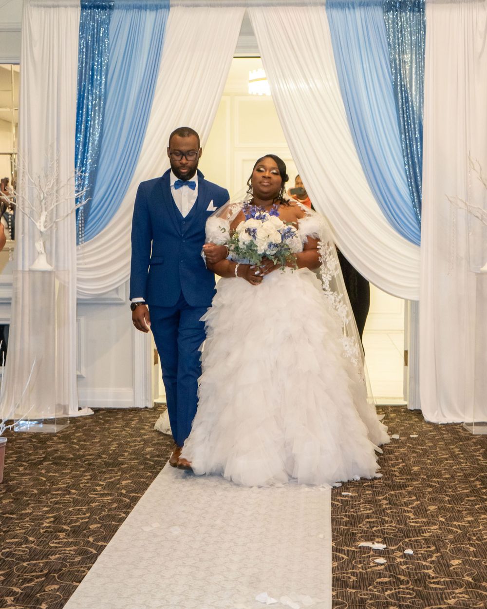 Couple walking down white aisle runner under elegant white and blue draped ceremony backdrop.