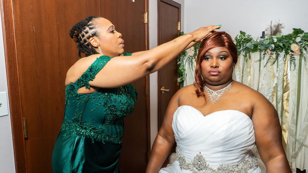 A bridesmaid in a green gown adjusts the hair of a bride in a white strapless wedding dress before the ceremony.